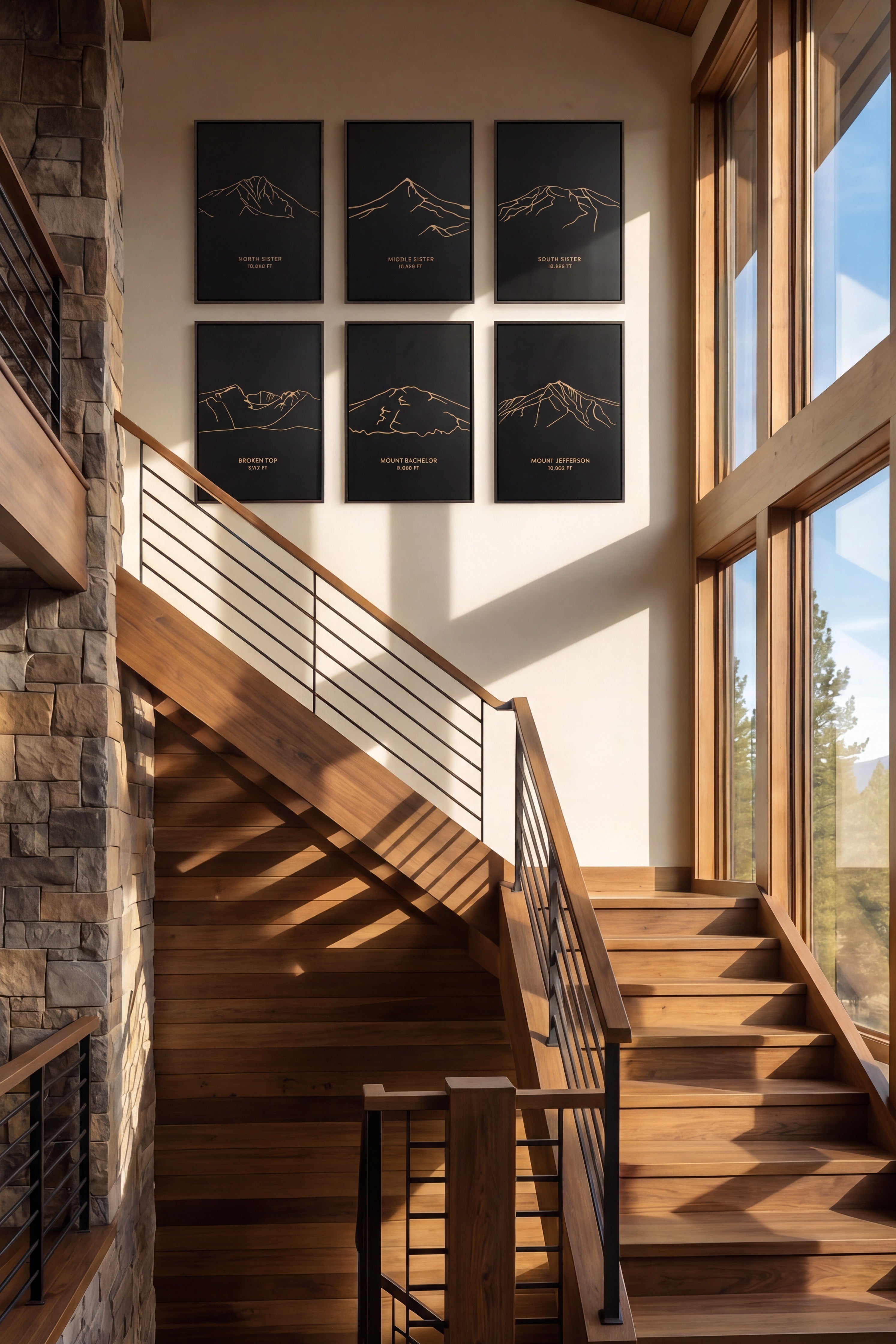 A modern wooden staircase in a rustic mountain-style home, with six framed black and white mountain line art prints on the wall above. Large windows on the right offer a view of pine trees and distant mountains.
